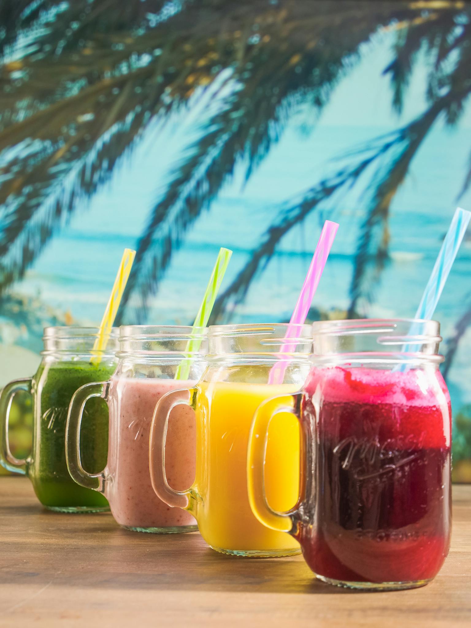 Colorful smoothies in mason jars with tropical backdrop, ideal for health and refreshment concepts.