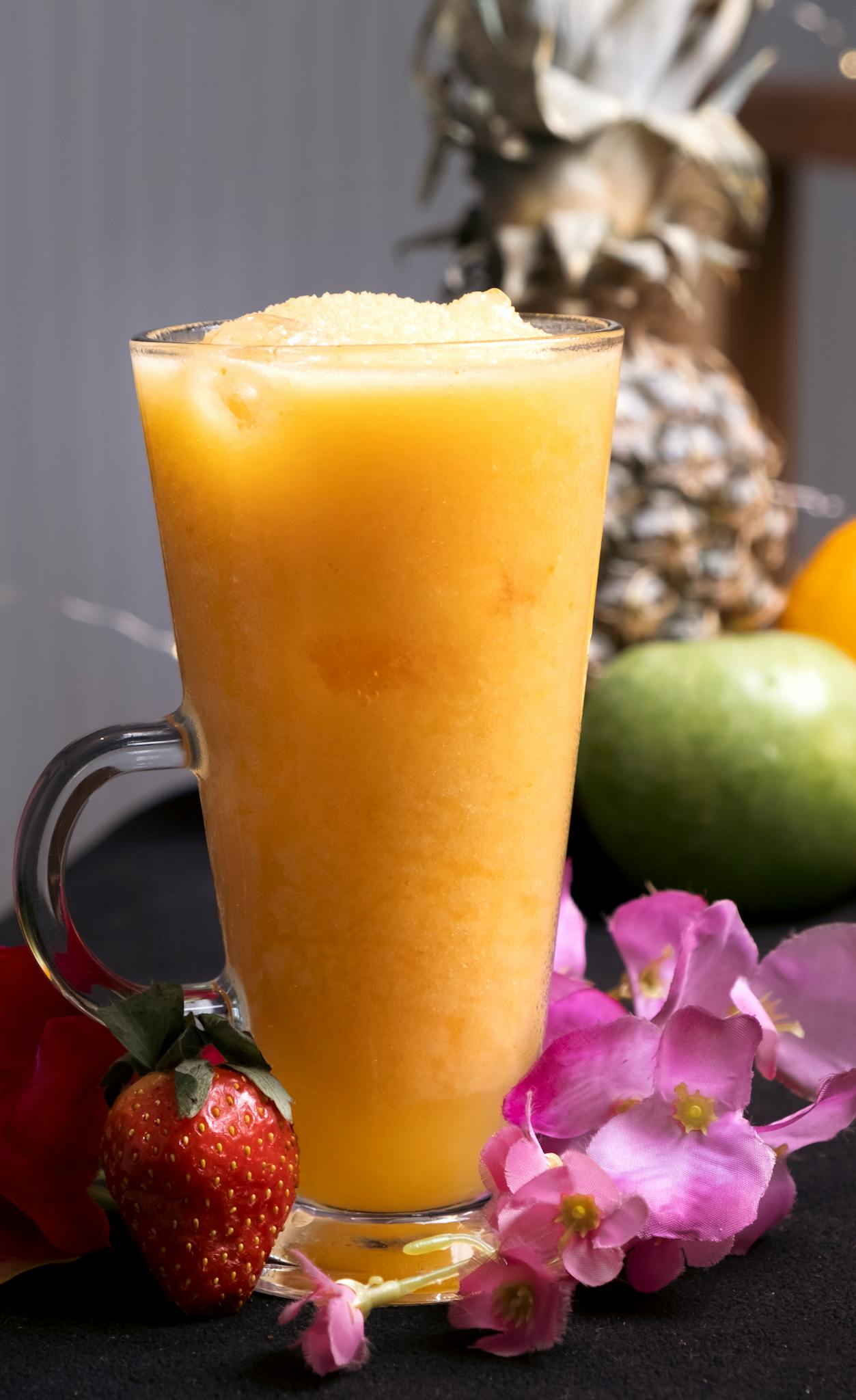 Delicious tropical fruit smoothie in a clear glass mug surrounded by fresh fruits and flowers.